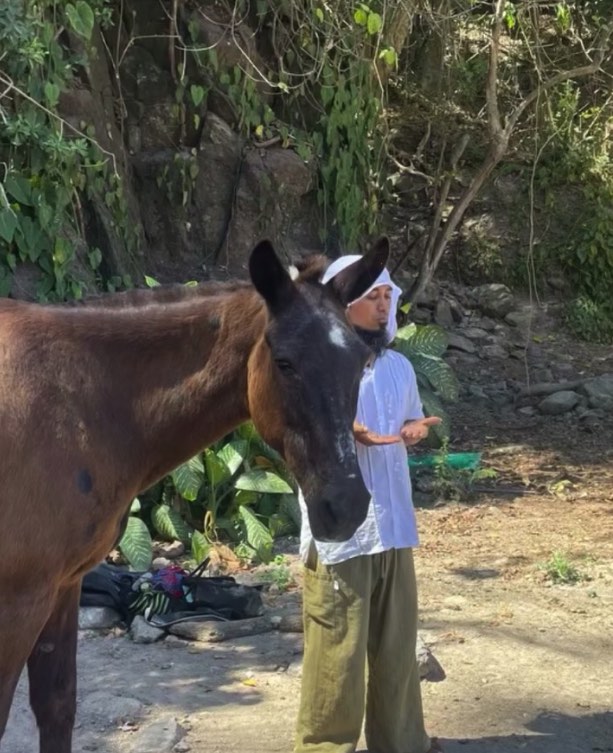 Una mujer de blanco, con las manos abiertas, en pie junto a un caballo oscuro